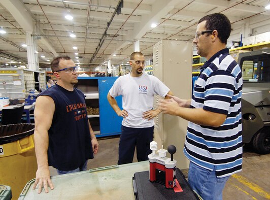 Bioenvironmental Engineering Flight member John Sharp explains a portable Wet Bulb Globe Temperature measuring device to 564th Aircraft Maintenance Group mechanics Jason Givens, left, and Pete Masura. The men work in the -135 kitting area of Bldg. 3705 where air conditioning is localized with large portable water drip cooling fans near their work stations and stepping outside the fan’s breeze can bring an instant sweat. The men start their days at 6:30 a.m. but with 23-plus days of triple-digit temperatures, mornings bring little relief. (Air Force photo by Margo Wright)