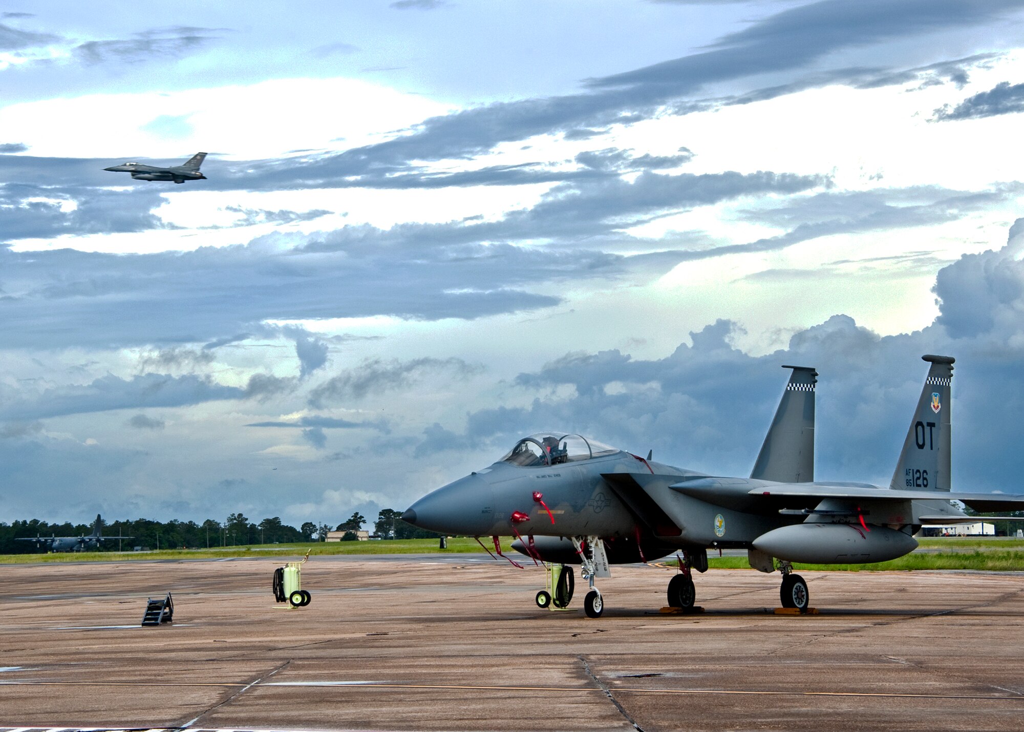 An F-15 sits on the Eglin Air Force Base, Fla., flightline as an F-16 Fighting Falcon soars over head and a C-130 taxis toward the runway, July 22.  These and other aircraft share Eglin’s flightline and runways with the Northwest Florida Regional Airport.   (U.S. Air Force photo/Samuel King Jr.)