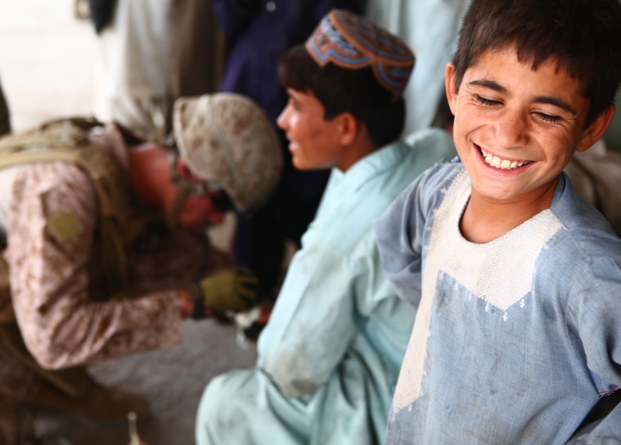 A young boy smiles for the camera while Petty Officer 3rd Class Brandon Dixon, a Navy corpsman with Guard Force, 1st Battalion, 6th Marine Regiment, treats a teenager's foot injury during a patrol here, July 29. The Marines and sailors of 1/6 took part in the initial offensive to loosen the insurgents' grip here, and the battalion has now returned, giving many within the unit a chance to see progress since their last deployment.
