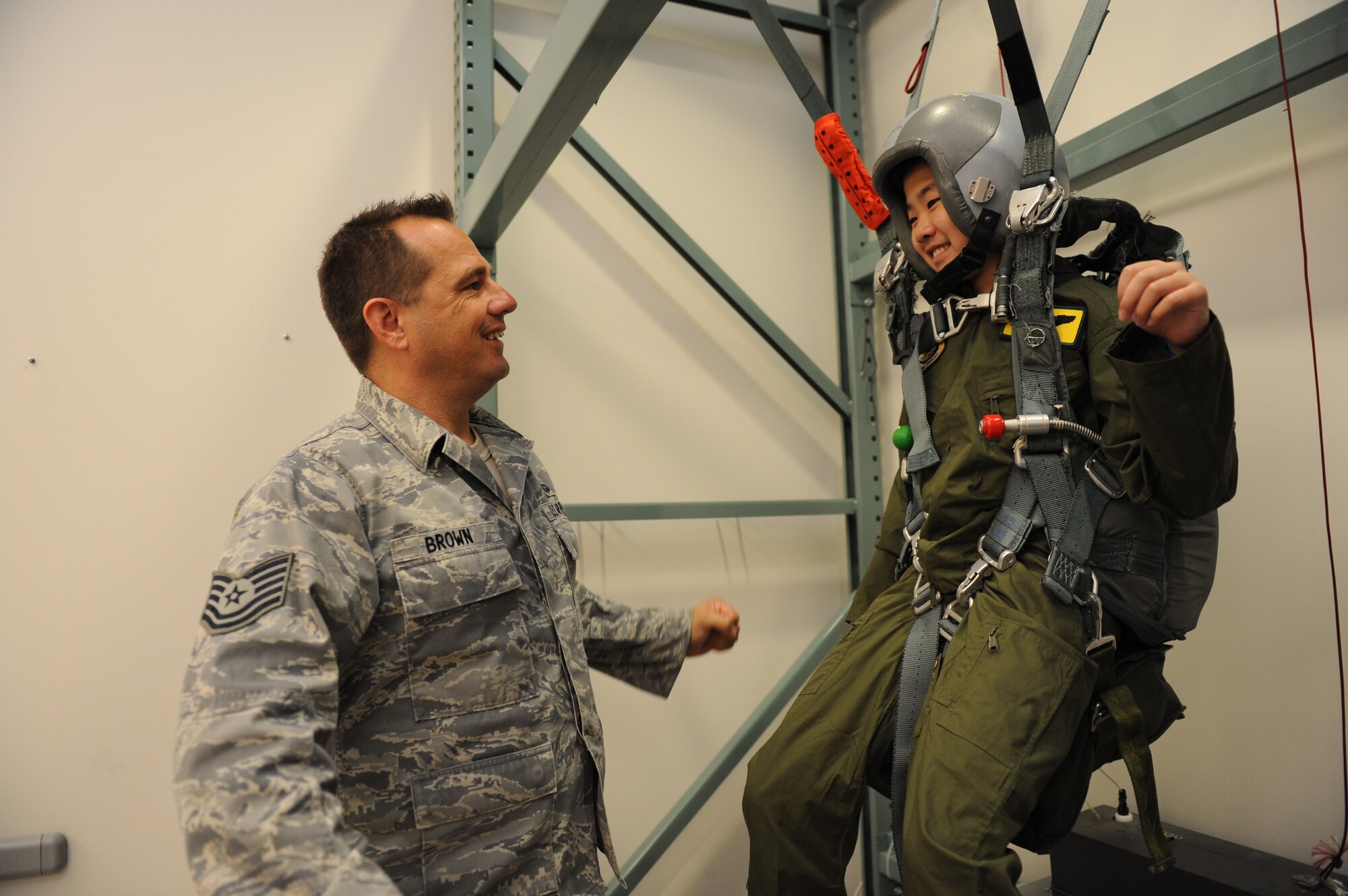 Tech. Sgt. Sherwood Brown, SERE specialist, assists Cody Sugai into a parachute training harness during a Pilot for the Day showcase of the squadrons on base June 10. Cody Sugai is this quarters "Pilot for a Day".  Cody is recovering from strokes caused by arteriovenous malformation and was nominated as the PFAD by Kapi'olani Medical Center in partnership with the 15th Wing. (U.S. Air Force photo/Senior Airman Lauren Main)