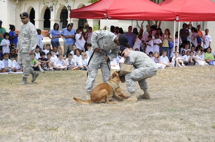 Senior Airman Kevin Banks, 902nd Security Forces Squadron military working dog handler, gives his K-9 partner, CCharlie, a command to release the training sleeve worn by Staff Sgt. Taylor Rogal, 902nd Security Forces Squadron military working dog trainer, during a demonstration at Operation Families Learning about Global Support on June 10. (US Air Force photo/A1C Precious Yett)
