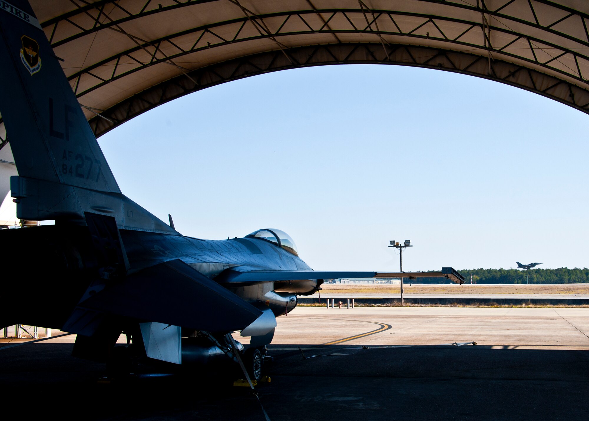 An F-16 from Luke Air Force Base, Ariz., waits for action while another Fighting Falcon performs a touch-and-go at Eglin's runway March 18.  The F-16s were brought here to provide familiarization training to the 33rd Fighter Wing pilots prior to the arrival of the F-35 and to establish a “battle rhythm” for the 33rd flightline operations.  Currently, Marines and Air Force pilots are completing daily sorties.  (U.S. Air Force photo/Samuel King Jr.)