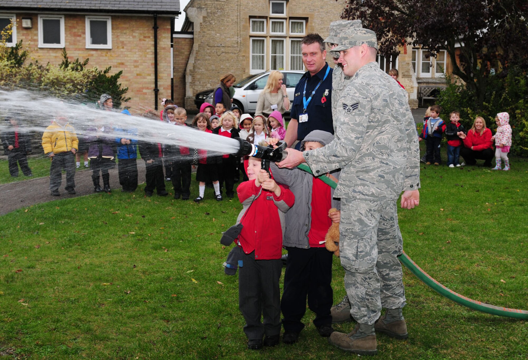 WEST ROW, England -- Senior Airman Shayne Anglin (front) and Staff Sgt.  Kyle Dulin, both 100th Civil Engineer Squadron Fire Department, and Watch Manager Ken Cross, 423rd Civil Engineer Squadron Fire Department, RAF Alconbury, assist children from West Row Primary School as they take turns holding a fire hose and spray water onto a grassy area at the school. The firefighters visited the school to foster continual good relations with the local community. (U.S. Air Force photo/Karen Abeyasekere)