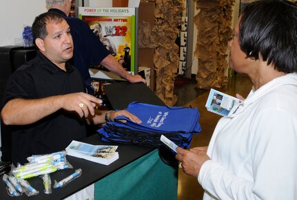 Juan Guerra, 902 Mission Support Group, Civil Engineering Environmental Flight, gives Dona Fair products made from recycled material as part of America Recylces Day, Nov. 15. Members of the Randolph community had an opportunity to learn more about recycling and to visit the recycling kiosk set up inside the Base Exchange with free promotional items made from recycled material. (U.S. Air Force Photo by Don Lindsey)