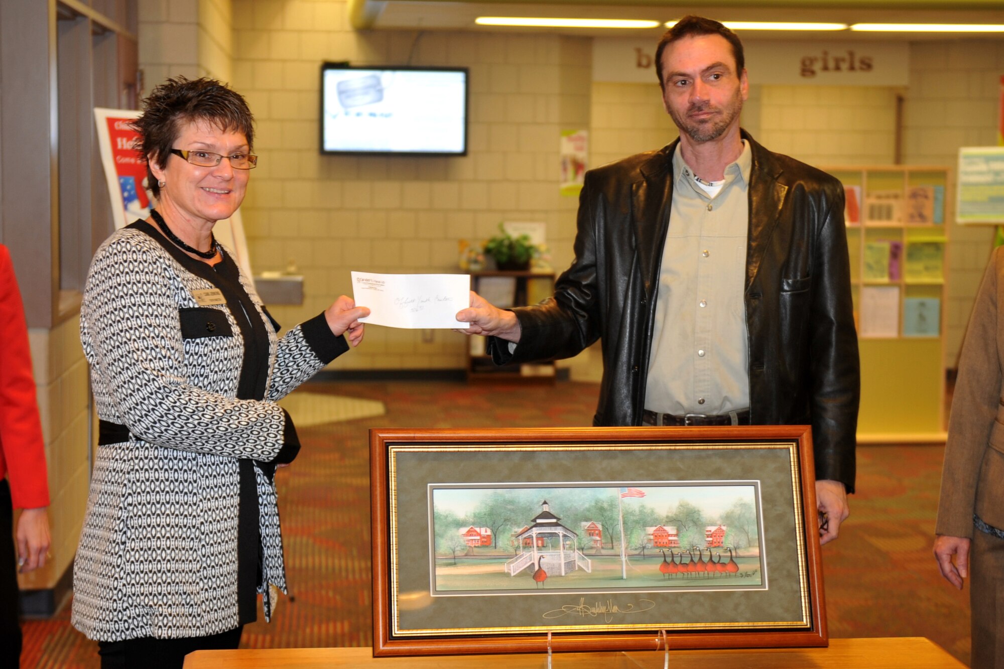 Jeff Bosiljevac, Ginger's Hang-up Framing store employee, presents a donation on behalf of artist P. Buckley Moss to Camela Jenkins, youth programs director, at the Offutt Youth Center on Offutt Air Force Base, Neb. Nov. 10. The artist donated money from the Offutt Parade Grounds print proceeds to benefit children of military members. (U.S. Air Force photo by Jeff W. Gates/Released)