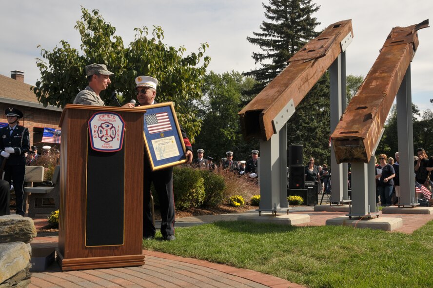 Col. Peter Siana presents a certificate and flag to the Enfield Fire Department during a ceremony commemorating the 10th anniversary of the terrorist attacks of 9/11, in Enfield, Conn., Sept. 11, 2011. The flag is significant in that it was flown in a Connecticut Air National Guard A-10 Thunderbolt II Aircraft during three tours of duty in Iraq and was airborne over Iraq at the exact moment of the attacks on our nation. To the right are two pieces of twisted steel salvaged from Ground Zero. (U.S. Air Force photo by Airman 1st Class Emmanuel Santiago)