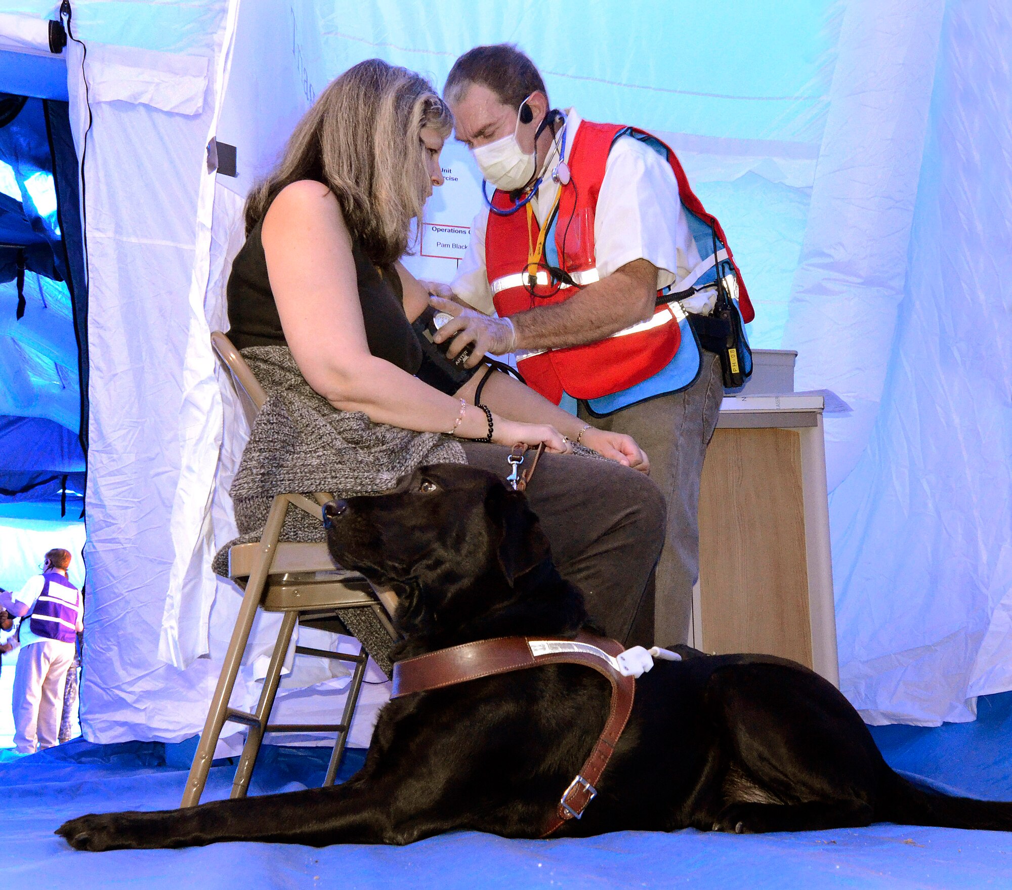 A Georgia State Defense Force doctor takes Rita Harrison's blood pressure while her guide dog sits-by patiently during a mass casualty exercise at Dobbins Air Reserve Base, Oct. 8. The Cobb & Douglas Public Health office ran the mass casualty exercise using their Mobile Surge Unit to help prepare local civilian medical personnel for a mass casualty event.   (U.S. Air Force photo/ Brad Fallin)