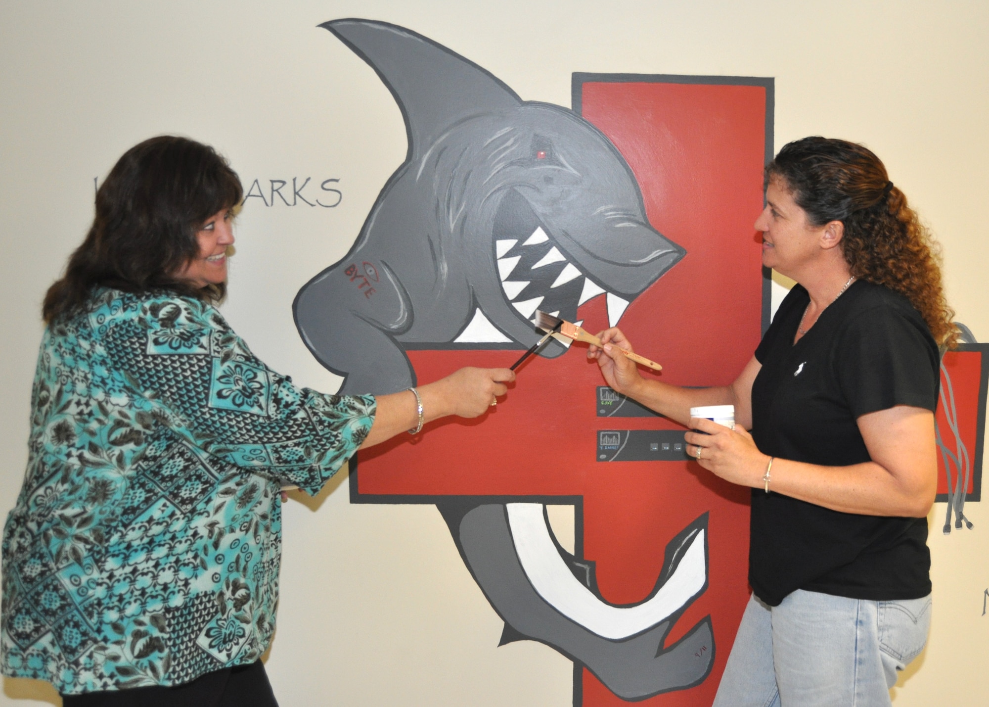 Tammy Raines, left, and Christine Roy, 81st Medical Support Squadron Medical information management flight, Keesler Air Force Base, Biloxi, Miss., cross paint brushes in front of the “LAN Shark” logo they created for the flight. They developed the mascot after flight commander Maj. Reginald Sennie suggested the unit could use something that might be incorporated into things like awards to enhance unit morale. They worked on the image over two non-training Fridays, completing it on Sept. 11, inscribing “9/11” within the shark’s tail. In addition to the words “LAN SHARK,” with LAN standing for “local area network,” the logo includes the term “NO LIMITS” — the letters “IMIT” representing “information management information technology.”  (U.S. Air Force photo by Steve Pivnick)