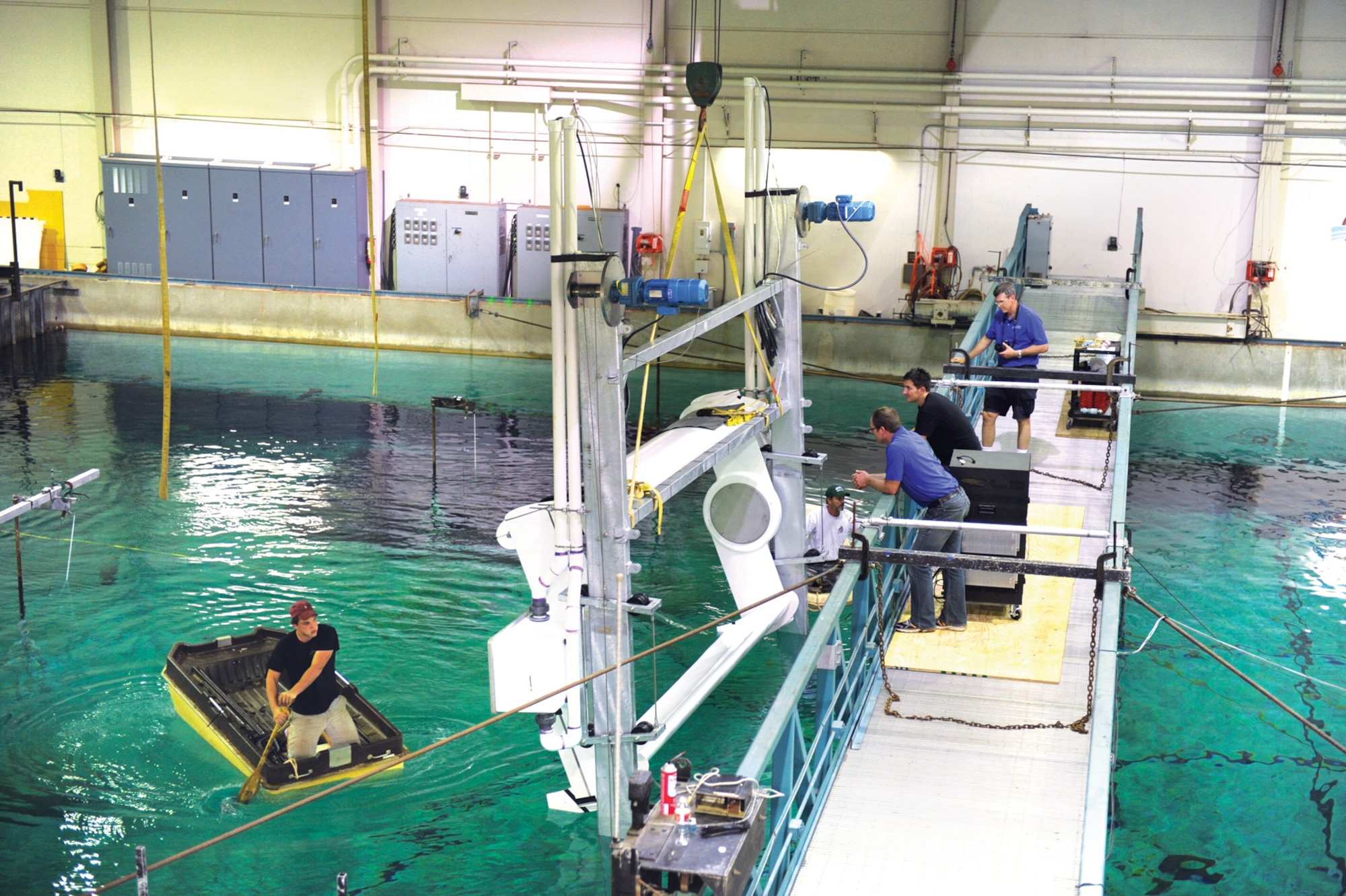 A technician at the Offshore Technology Research Center positions his boat to remove the brackets which secured the wave energy converter’s blades after the 5-ton crane lifted it into position over the test pool, while other members of the Academy’s wave energy team and testing center staff make preparations to lower the converter into the testing pool.  (U.S. Air Force Photo/John Van Winkle)