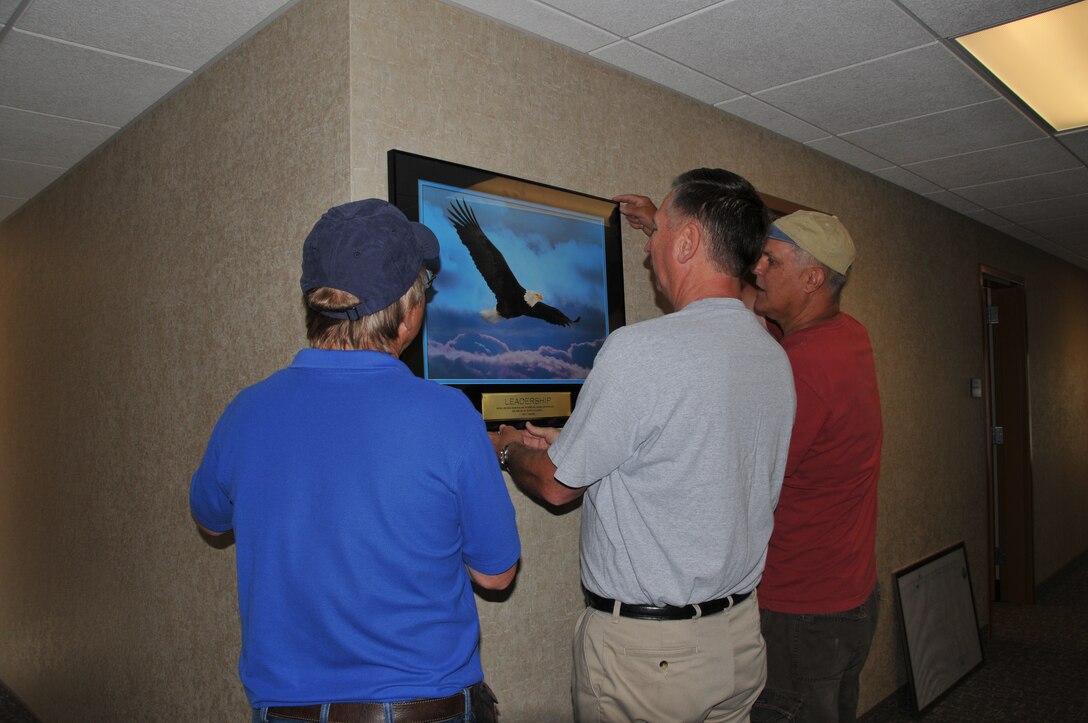 Retired Senior Master Sgt. Mark Giskass, retired Col. Fred Fairhurst and retired Chief Master Sgt. Dana Darko hang a lithograph on the wall.
 (U.S. Air Force photo by Senior Master Sgt. Eric Peterson.)