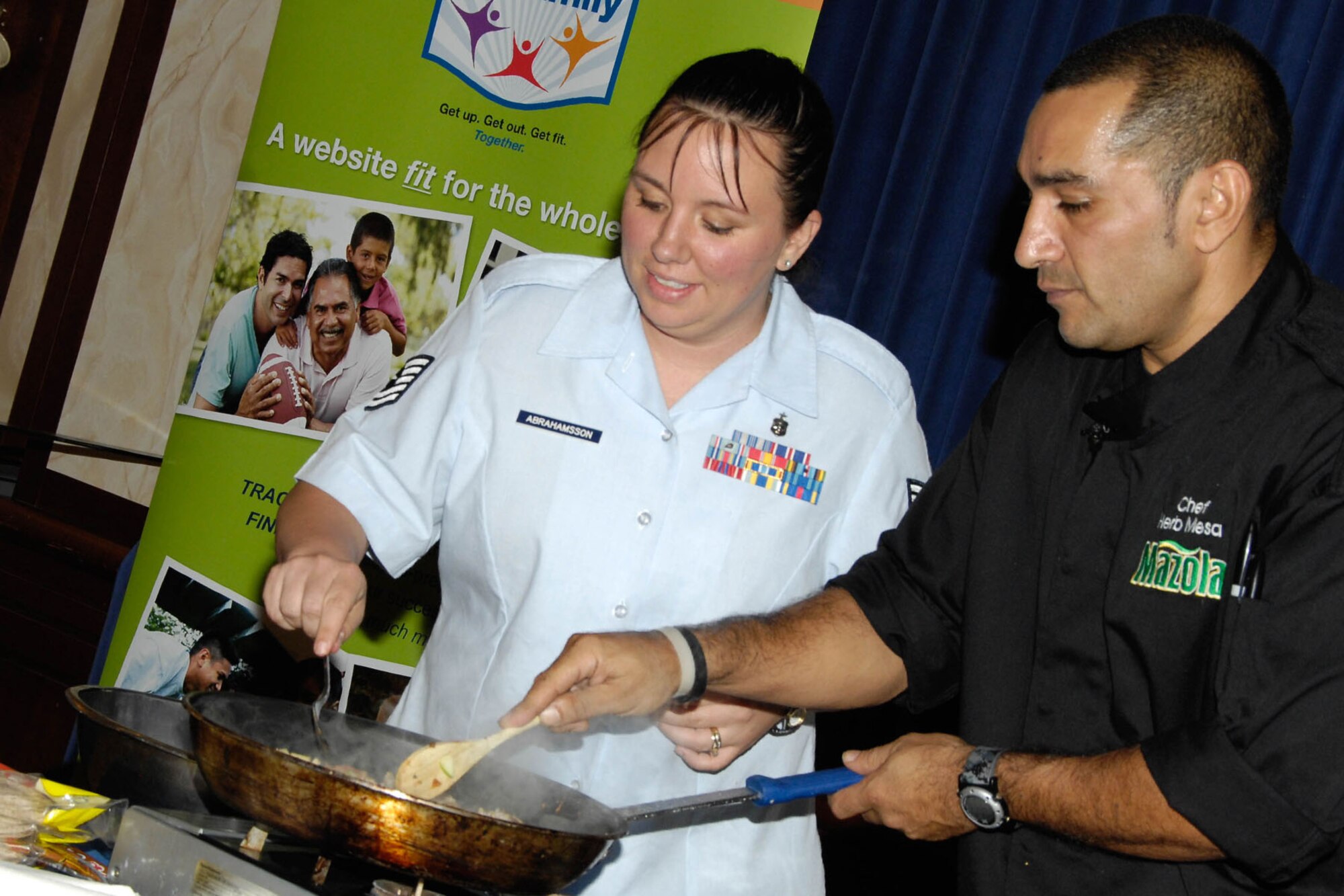 HANSCOM AIR FORCE BASE, Mass. – Tech. Sgt. Michelle Abrahamsson, winner of the Air Force Fit Family "Cook It Up" Healthy Recipe Contest, assists celebrity chef Herb Mesa during a cooking demonstration in the Den at the Minuteman Commons grand opening Sept. 8. Just before the demonstration, Chef Mesa presented Sergeant Abrahamsson with an award for her winning recipe, Apple Turkey Picadillo. (U.S. Air Force photo by Linda LaBonte Britt)
