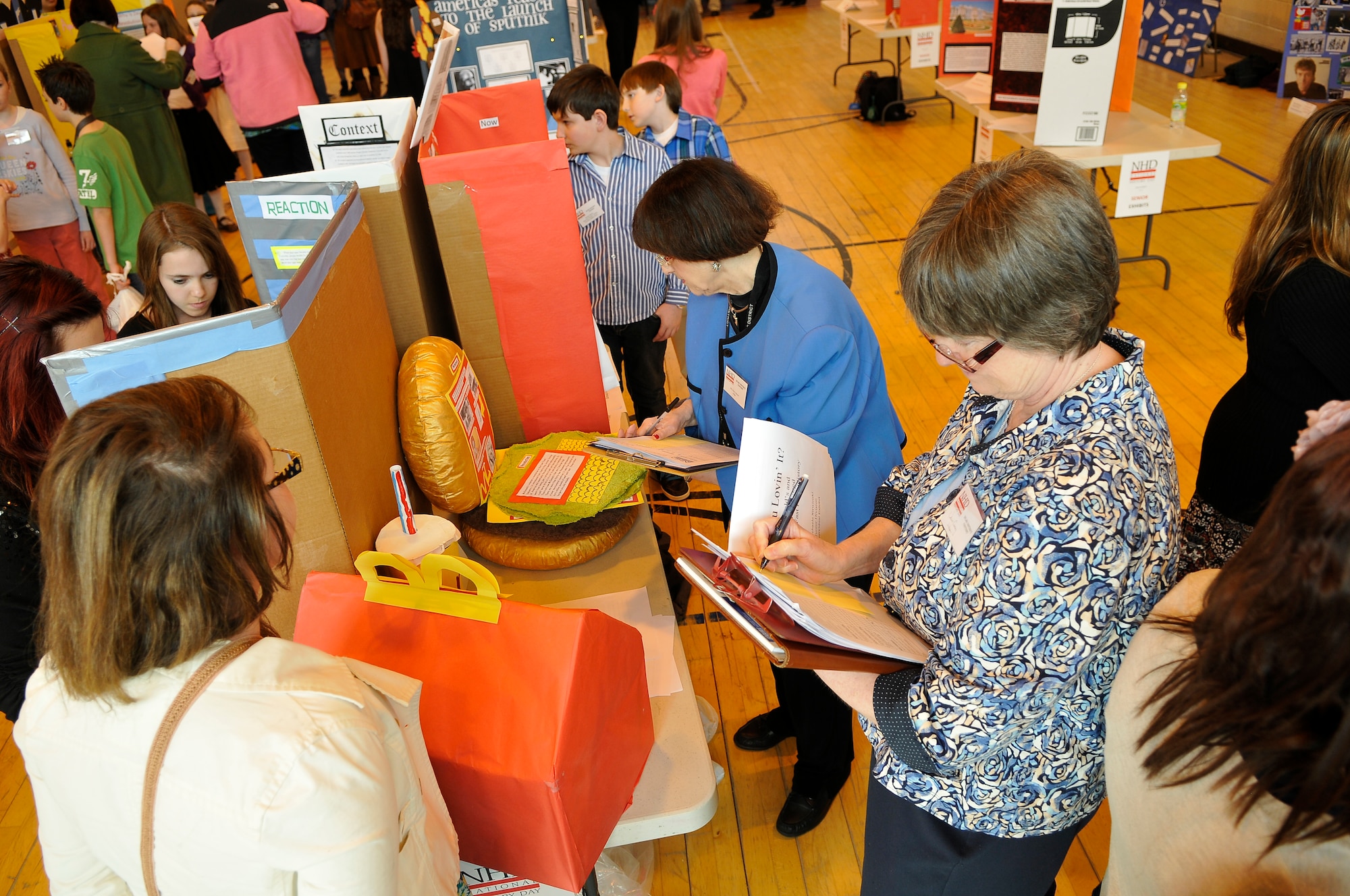 ROYAL AIR FORCE LAKENHEATH, England -- Judges take notes on one of 45 exhibits during the National History Day contest at the Lakenheath High School gym April 3, 2012.  National History Day is a national academic program for students grades 6 through 12. The competition benefits students by promoting reading, writing, research skills and building connections with fellow students interested in history. (U.S. Air Force photo by Staff Sgt. Connor Estes)
