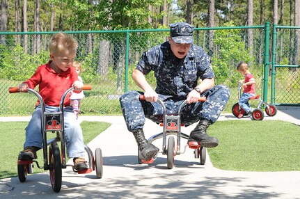 Three-year old Logan Sorensen passes Petty Officer 3rd Class Steven Swan on his tricycle during play time at the Child Development Center at Joint Base Charleston – Weapons Station April, 11. Swan, a Machinist’s Mate from Rockmart, Ga., is working at the CDC while he waits on his next set of orders. Logan is the son of Chief Petty Officer Shawn Sorensen, Naval Support Activity command career counselor. (U.S. Navy photo/Petty Officer 1st Class Jennifer Hudson)