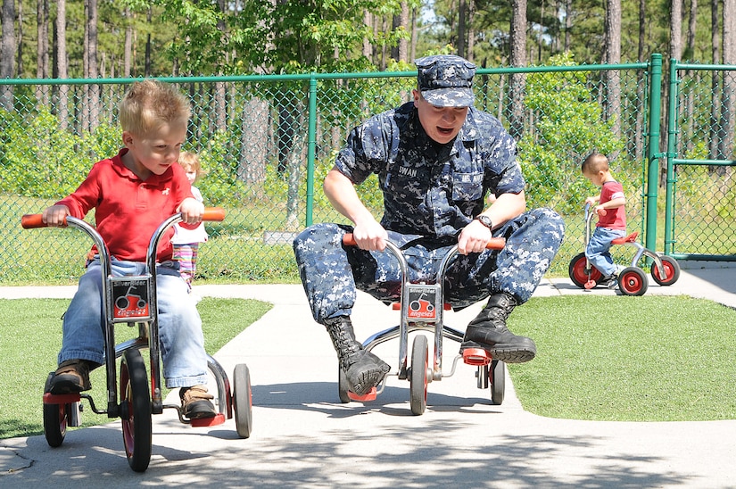 Three-year old Logan Sorensen passes Petty Officer 3rd Class Steven Swan on his tricycle during play time at the Child Development Center at Joint Base Charleston – Weapons Station April, 11. Swan, a Machinist’s Mate from Rockmart, Ga., is working at the CDC while he waits on his next set of orders. Logan is the son of Chief Petty Officer Shawn Sorensen, Naval Support Activity command career counselor. (U.S. Navy photo/Petty Officer 1st Class Jennifer Hudson)