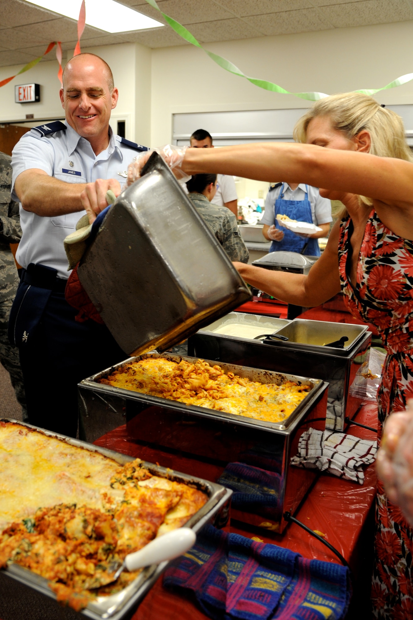 Col. Darren James, left, the 71st Flying Training Wing commander, helps fill the serving line at the July Singles’ International Gourmet Meal Opportunity held July 30 in the Community Chapel Activity Center at Vance Air Force Base, Okla. Spooning food from the tray is Dr. Debo’rah Merritt, whose husband is Lt. Col. Michael Merritt, the 71st FTW vice commander. SIGMO is a free home-cooked meal offered monthly to single Airmen and the families of deployed Airmen at Vance AFB. (U.S. Air Force photo/ Airman 1st Class Frank Casciotta)