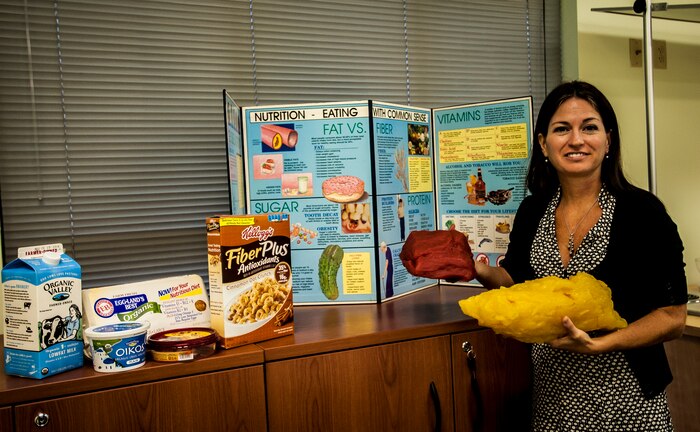 Wendy Wyatt, Health and Wellness Center dietician, compares five pounds of fat to five pounds of muscle. Behind Wyatt is a healthy eating display she uses when teaching nutritional classes. Wyatt is a registered dietician who recently joined the HAWC team. The HAWC’s mission is to cultivate a fit and healthy force. (U.S. Air Force photo by Senior Airman Dennis Sloan/Released)