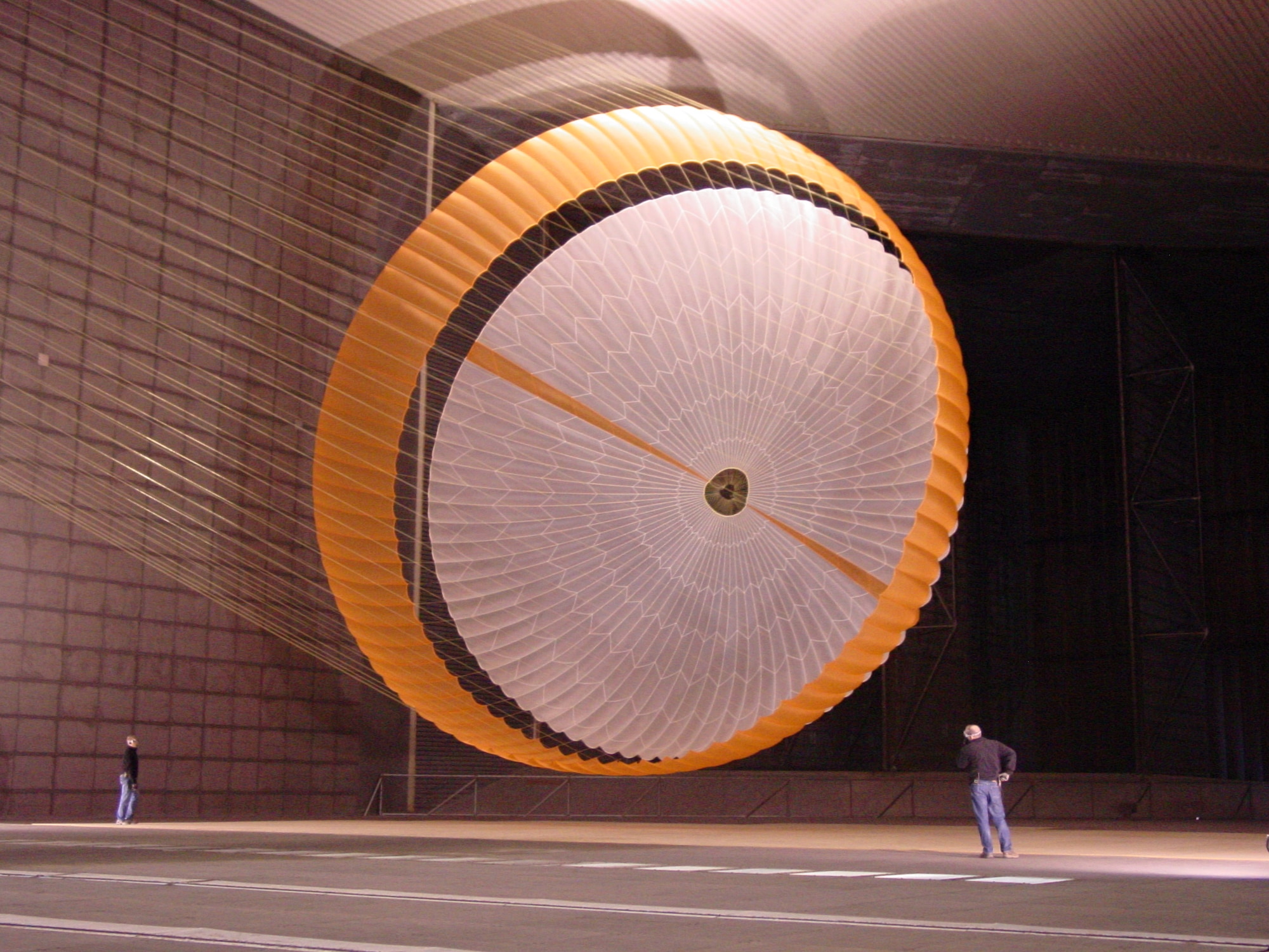 In this image, two engineers are dwarfed by NASA's Mars Science Laboratory's parachute, which holds more air than a 3,000-square-foot house and is designed to survive loads in excess of 36,000 kilograms (80,000 pounds). The parachute, built by Pioneer Aerospace, South Windsor, Conn., has 80 suspension lines, measures more than 65 feet in length, and opens to a diameter of nearly 55 feet. It is the largest disk-gap-band parachute ever built and is shown here inflated in the test section with only about 12.5 feet of clearance to both the floor and ceiling of the world’s largest wind tunnel at National Full-Scale Aerodynamics Complex. The parachute is attached to a launch arm mounted on a swivel-base that allows the test item to pitch and yaw under simulated conditions of subsonic entry into the Martian atmosphere. (Photo courtesy of NASA/JPL and Pioneer Aerospace Corp.)