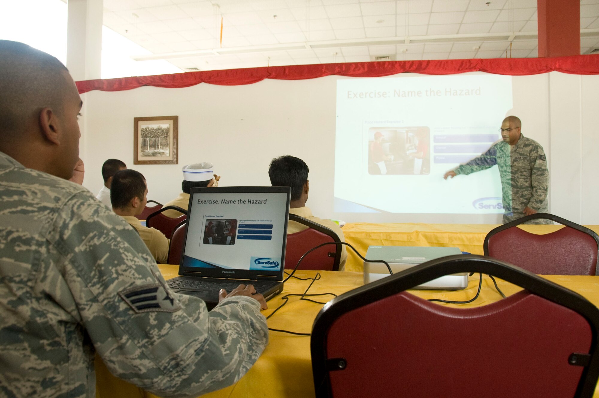 Senior Airman Joel Varghese, (left) 379th Expeditionary Medical Group Public Health force health program manager, works the PowerPoint slides as Staff Sgt. Ricardo Lemos, 379th EMDG Public Health operations manager, trains dining facility food handlers Aug. 6, 2012 on the importance of proper food handling techniques, good personal hygiene, hand washing, proper use of gloves and keeping their hair covered. Varghese and Lemos are Public Health Airmen whose primary job is to keep deployed Airmen healthy in Southwest Asia. (U.S. Air Force photo/Senior Airman Bryan Swink)