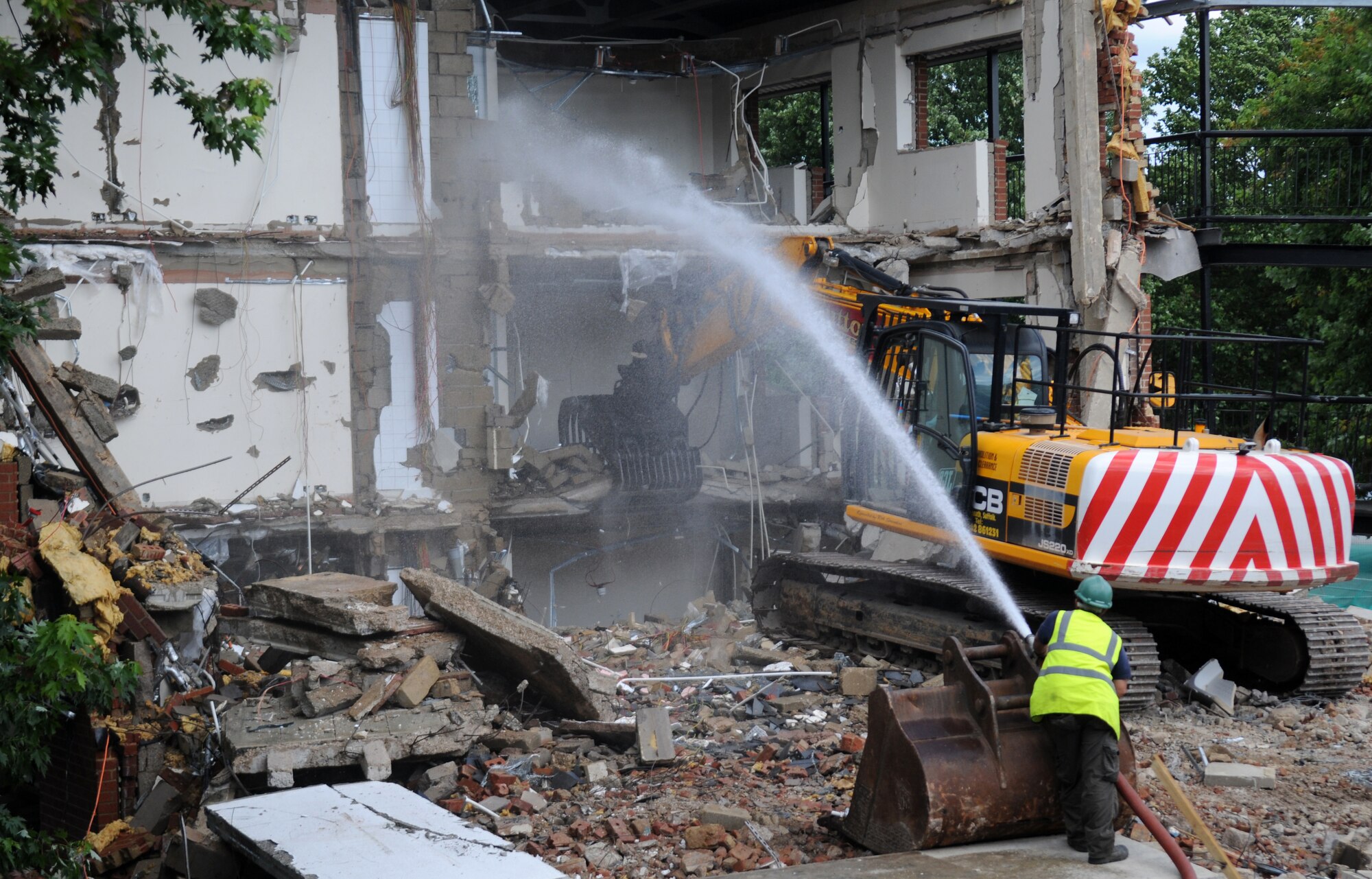 RAF MILDENHALL, England – Neil Drike, a contractor, sprays Dorm 214 with water as Rob Saunvers, JCB Tracked Excavator JS220 Long Reach operator and contractor, demolishes the building August 17, 2012, at RAF Mildenhall. The dorm demolishment project is scheduled to be completed by the end of September 2012 and is expected to cost $389,000. (U.S. Air Force photo/Senior Airman Rachel Waller)       