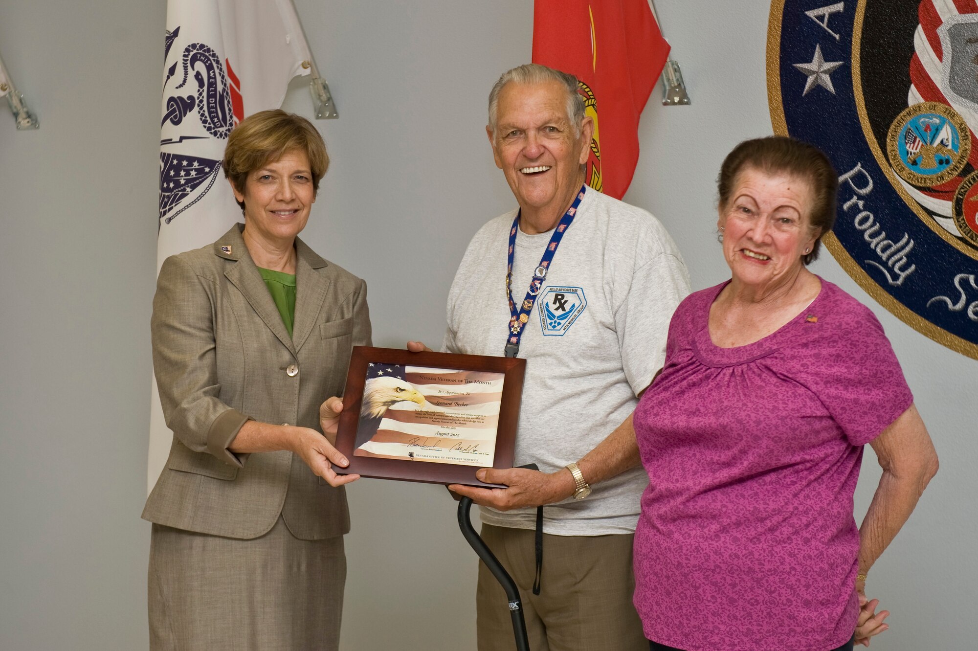 Katherine Miller, deputy executive director of the Nevada Office of Veterans Services presents the Veteran Volunteer of the Month Award to Leonard Becker, accompanied by his wife Elva Becker, Aug. 23, 2012, at Nellis Air Force Base, Nev. Mr. Becker helped the Nellis AFB Pharmacy return outdated prescriptions which, in turn, helped the facility save $40k annually. (U.S. Air Force photo by Airman 1st Class Daniel Hughes)