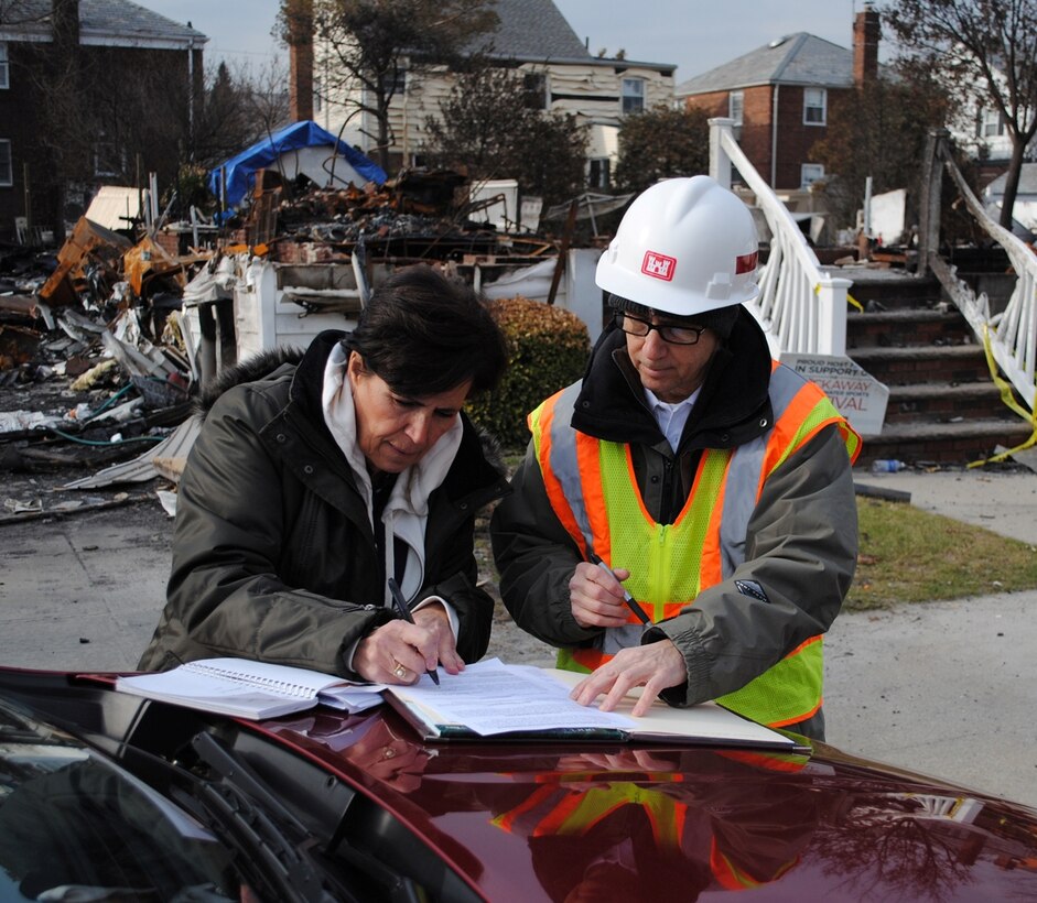 Martha Militano, a resident of the Rockaways in Queens, N.Y., signs a right-of-entry form, Nov. 30, authorizing the U.S. Army Corps of Engineers to remove the remains of her home, pictured behind her, burned to the ground during Hurricane Sandy. Assisting her is Leslie Williams, a real estate specialist with the Army Corps New York Recovery Field Office.