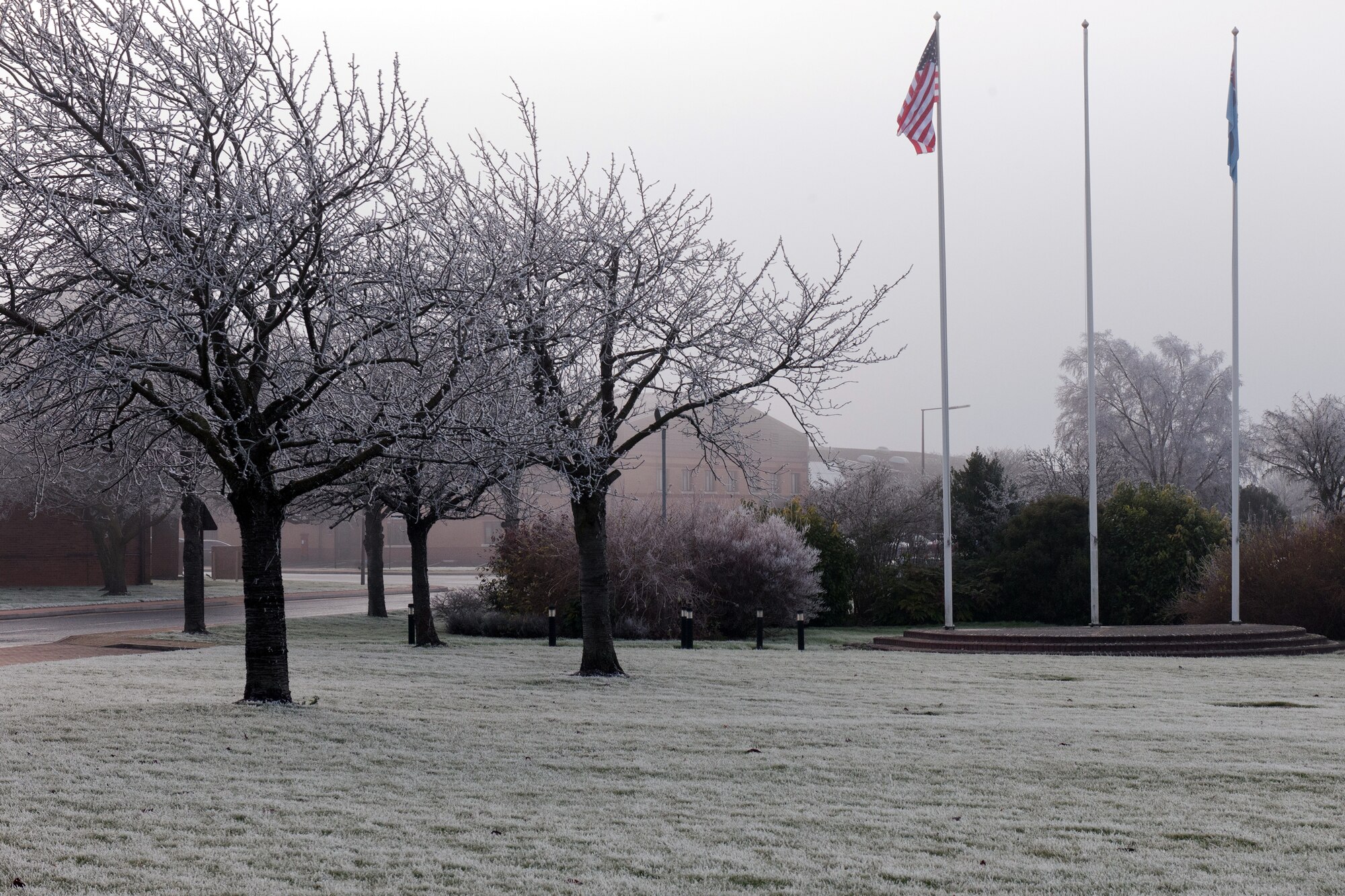 RAF ALCONBURY, United Kingdom – The area around the RAF Alconbury flagpoles is covered in frost, called hoar frost, Dec. 12. Hoar frost (also called radiation frost, hoarfrost or pruina) refers to the white ice crystals, loosely deposited on the ground or exposed objects, which form on cold clear nights when heat losses into the open skies cause objects to become colder than the surrounding air. The frost itself will be at or below the freezing temperature of water. The name hoar comes from an Old English adjective for showing signs of old age, and is used in this context in reference to the frost, which makes trees and bushes look like white hair. (U.S. Air Force photo by Staff Sgt. Brian Stives)