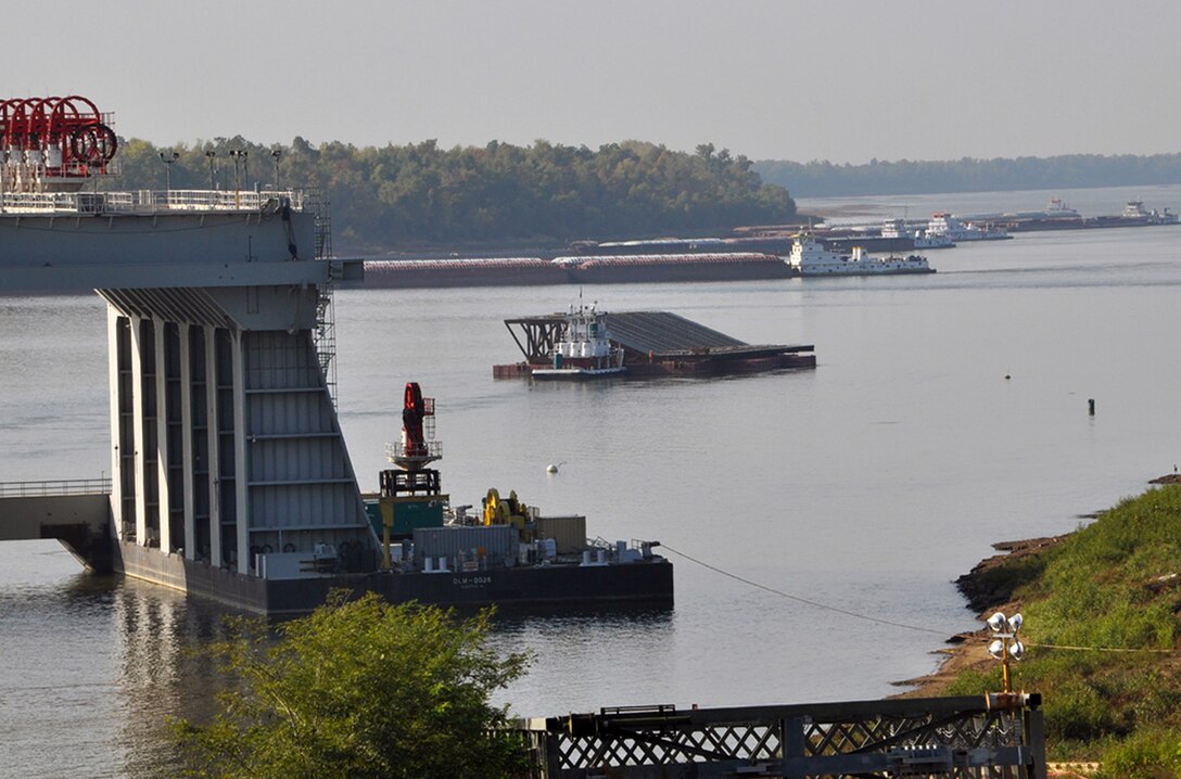 In the upper right, tens of thousands of tons of commodities on scores of barges wait in queue Oct. 16 in front of their tow for a turn to pass through Locks and Dam 53 on their way up the Ohio River. In the lower left, a partial view of the catamaran barge used to transport from the casting yard the giant concrete and steel dam shells and place them in the river. The giant monoliths will become part of the Olmsted Locks and Dam that will replace both antiquated locks and dams 52 and 53 and greatly reduce lockage times. 