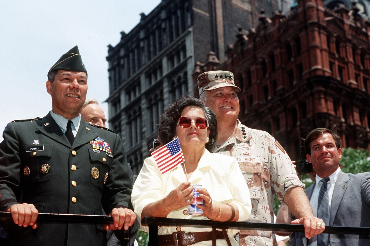 Two Army generals and a woman holding a flag ride on a float in a parade.