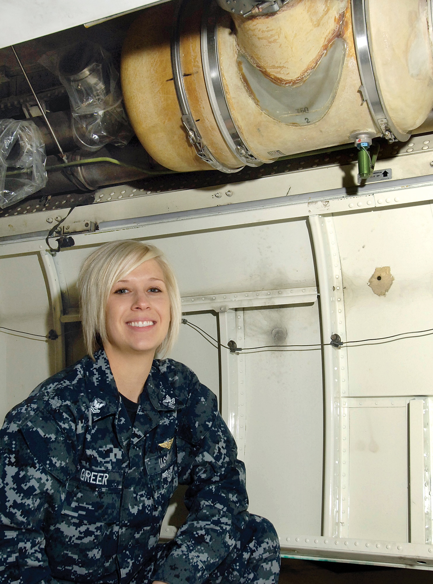 Petty Officer 2nd Class Brandi Greer, Fleet Air Reconnaissance Squadron FOUR, kneels by an E-6B Mercury jet at Tinker.  Weary after a 13-hour flight out-of-state and with only a small flashlight at night, the petty officer’s training and instinct helped her find a crack in the air cycle machine water separator input joint behind a bay panel under the belly of the plane. If undetected, the part could have failed bringing the jet down on its next mission.  For her action, Petty Officer Greer was named “Safety Pro of the Year” for the entire Navy wing here. (Air Force photo by Margo Wright)