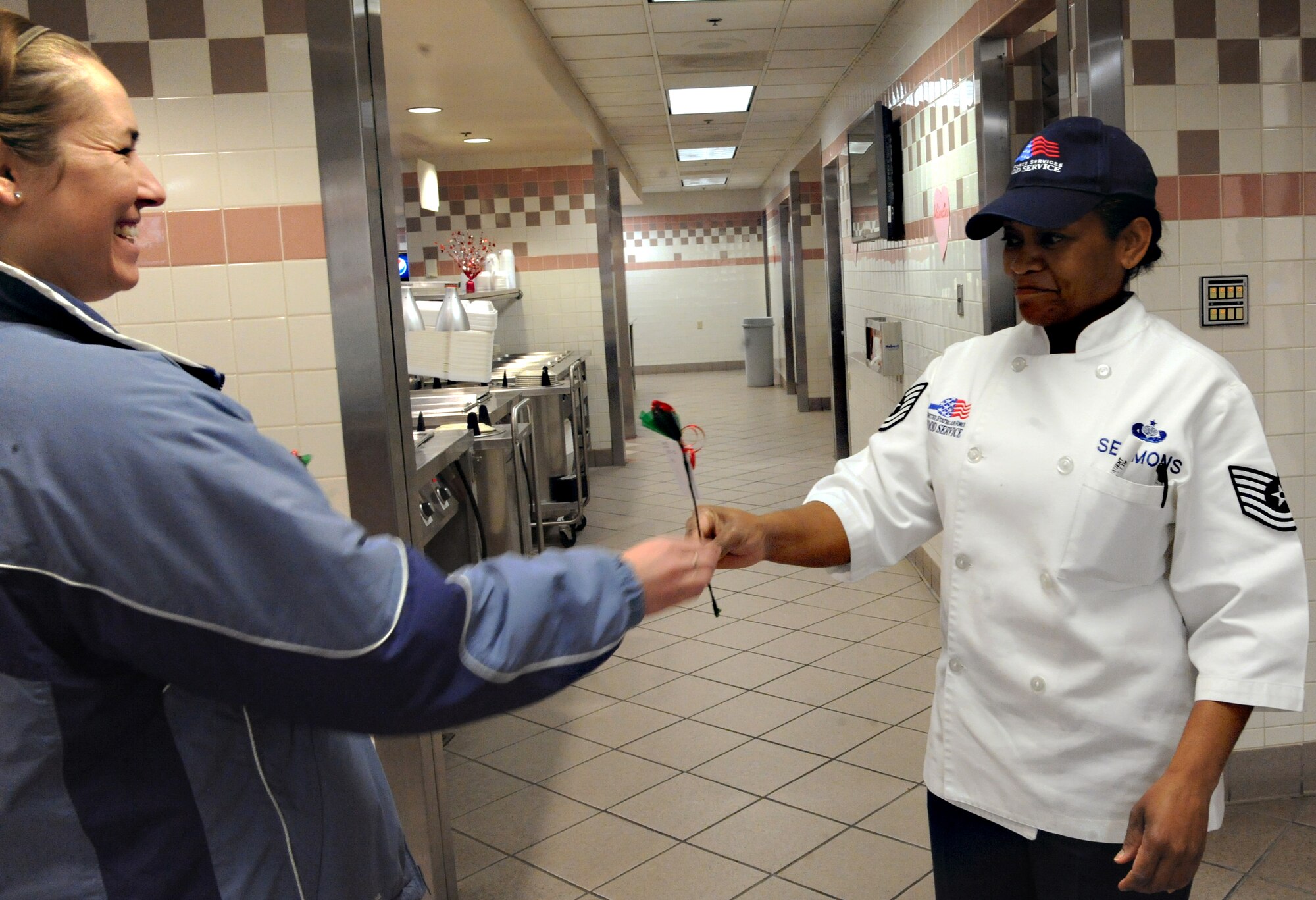 Tech. Sgt. Breezy Pruitt (left) presents Tech. Sgt. Areda Sermons (right), both members of the 319th Force Support Squadron, with a “Sweetheart Rose” from a secret admirer in the Airey Dining Facility on Feb. 14 on Grand Forks Air Force Base, N.D.  The Sweetheart Rose came from Operation Cupid which helped raise $291 for Network 56.  Network 56 is a professional organization with the intentions of reaching out to the community and making a difference in themselves and others. (U.S. Air Force Photo by Airman 1st Class Xavier Navarro) 