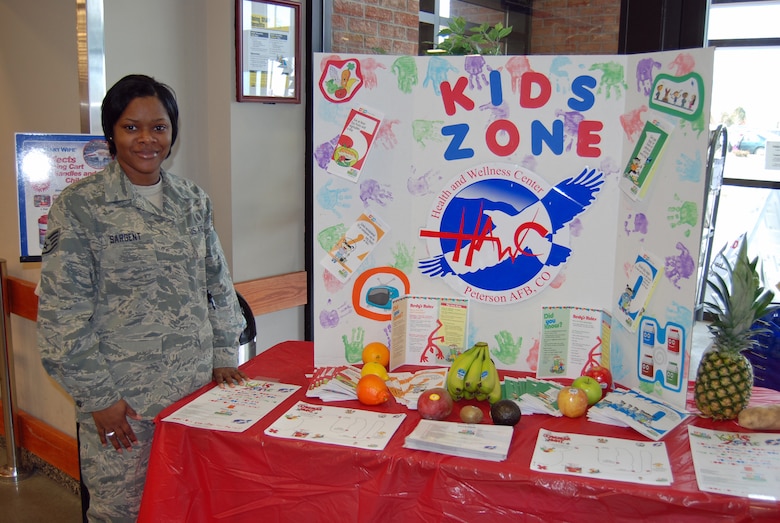 Staff Sgt. Ronneisha Sargent, Health and Wellness Center dietary therapist, stands by a display at the commissary to hand out a scavenger hunt and recipe cards to children. The scavenger hunt is part of the 5210 Let’s Go! program promoting five servings of fruits and vegetables, less than two hours of screen time, one hour of physical activity, and zero sugary drinks. Each week a different kid-friendly, healthy recipe will be available at the table. (U.S. Air Force photo/Lea Johnson)