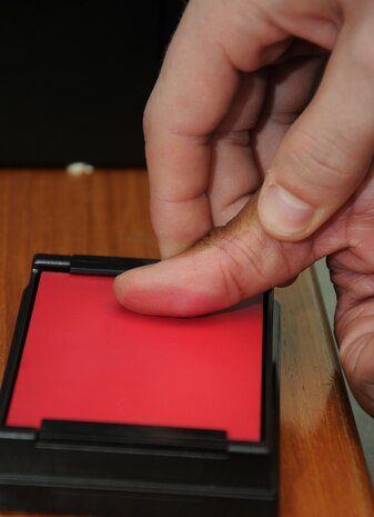 Petty Officer 2nd Class Eric Martin rolls Petty Officer 3rd Class Ryan Davis’ thumb on an inkless pad system before placing the print on the fingerprinting card at Joint Base Charleston – Weapons Station. Instead of using traditional black ink, the new system allows members to wipe away the red smear with little problem. (U.S. Navy photo/Petty Officer 1st Class Jennifer Hudson)