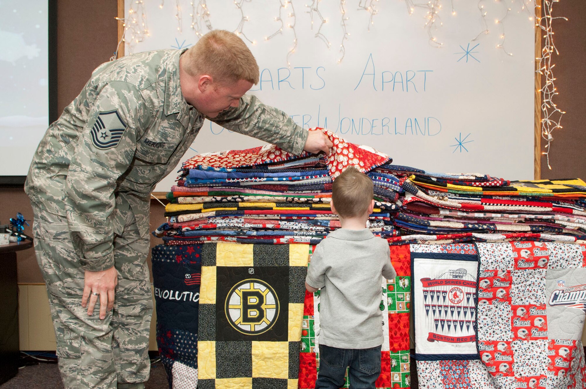 HANSCOM AIR FORCE BASE, Mass. – Master Sgt. Raymond Merring, Airman and Family Readiness Center, helps Joey DeNise, 3, choose a donated quilt made by Wrapped up in Sports during the Hearts Apart event at the A&FRC Jan. 19. Families of deployed military personnel receive the quilts by Wrapped up in Sports, who rally volunteer quilters to make and collect professional sports team-inspired quilts out of recycled sports apparel and  team fabrics. The Hearts Apart program offers support and activities for families that have loved ones deployed. (U.S. Air Force photo by Mark Wyatt)