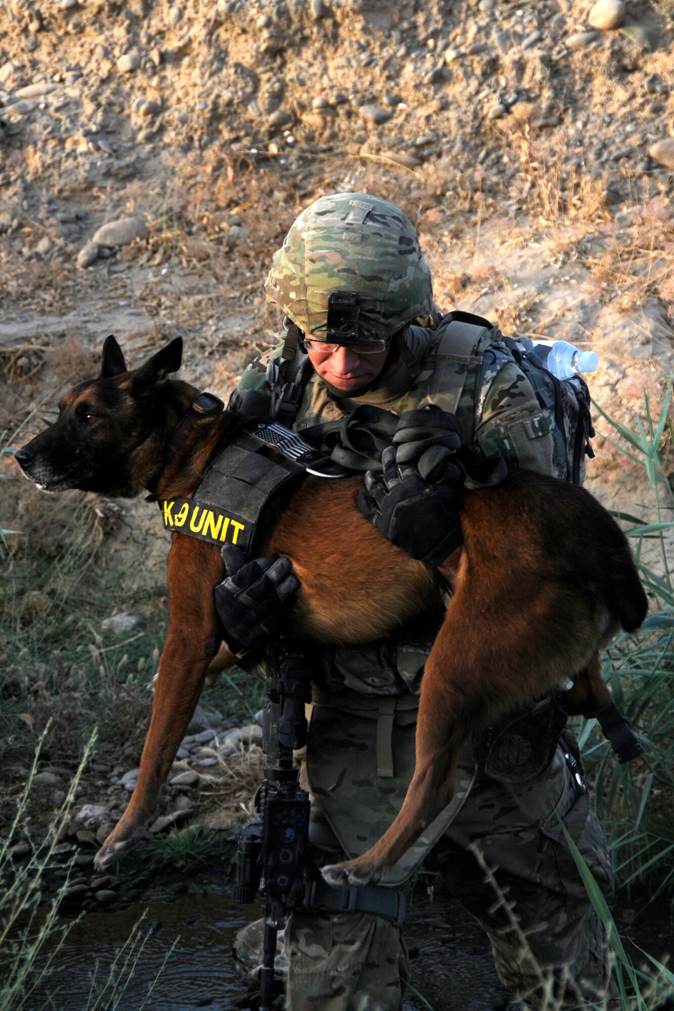 U.S. Air Force Staff Sgt. Chris Fall, a Military Working Dog handler, carries his dog, Glenn, through a ditch at Kandahar Airfield, Afghanistan on July 9, 2012. Glenn is a Patrol Explosive Detection dog and together they are working on their validation prior to deploying to a Forward Operating Base to work with ground forces seeking out IEDs on combat foot patrols. (U.S. Air Force photo/TSgt. Stephen Hudson)