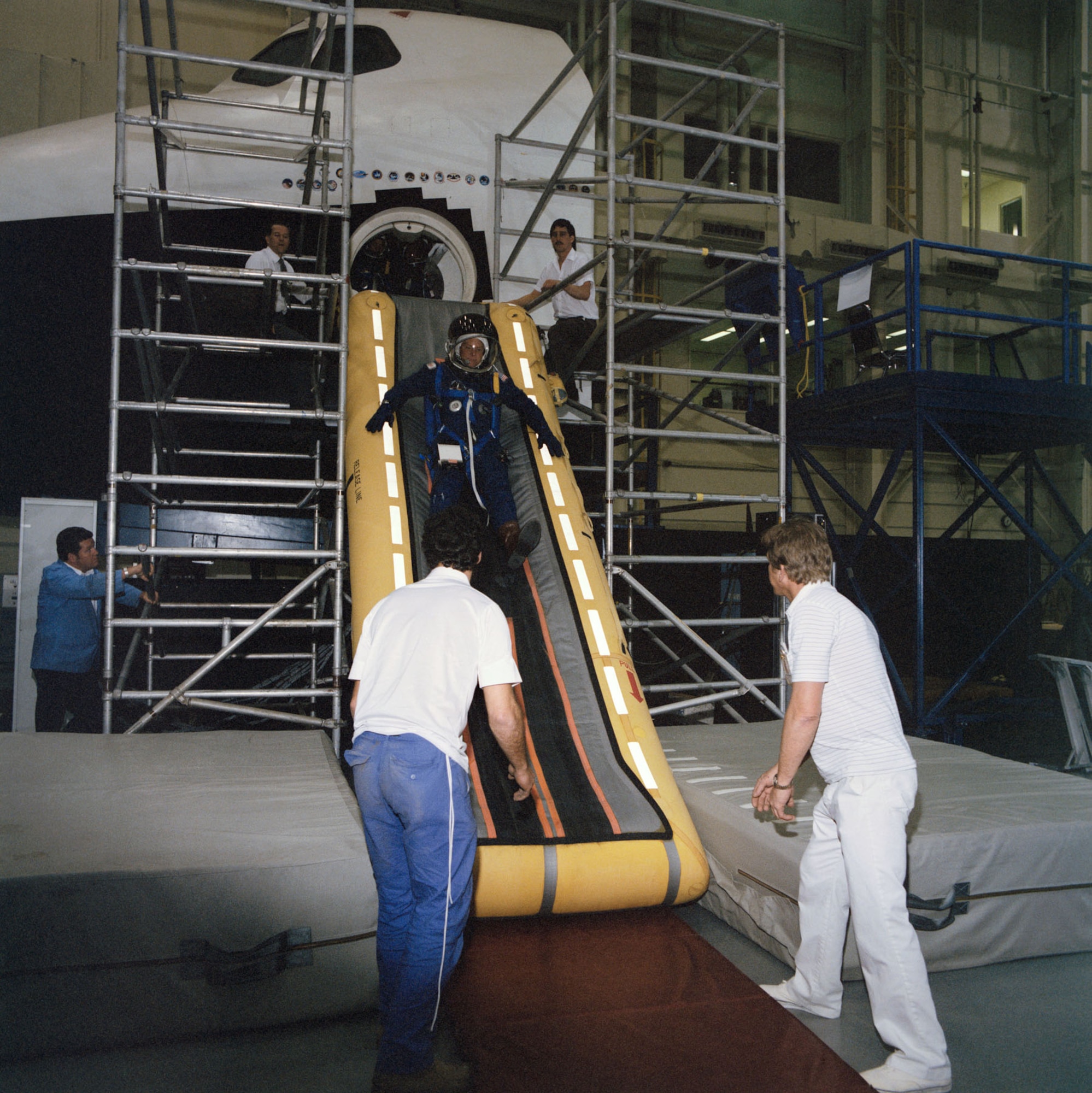 STS-26 Mission Specialist George D. Nelson trains in the Crew Compartment Trainer (CCT) located in Johnson Space Center's Shuttle Mockup and Integration Laboratory in March 1988. Nelson, wearing a partial pressure suit and helmet, exits the CCT via a slide inflated at the side hatch. Technicians at the bottom of the slide prepare to help Nelson to his feet as a second set of technicians observe the activity from scaffolding on either side of the open hatch. (NASA photo)