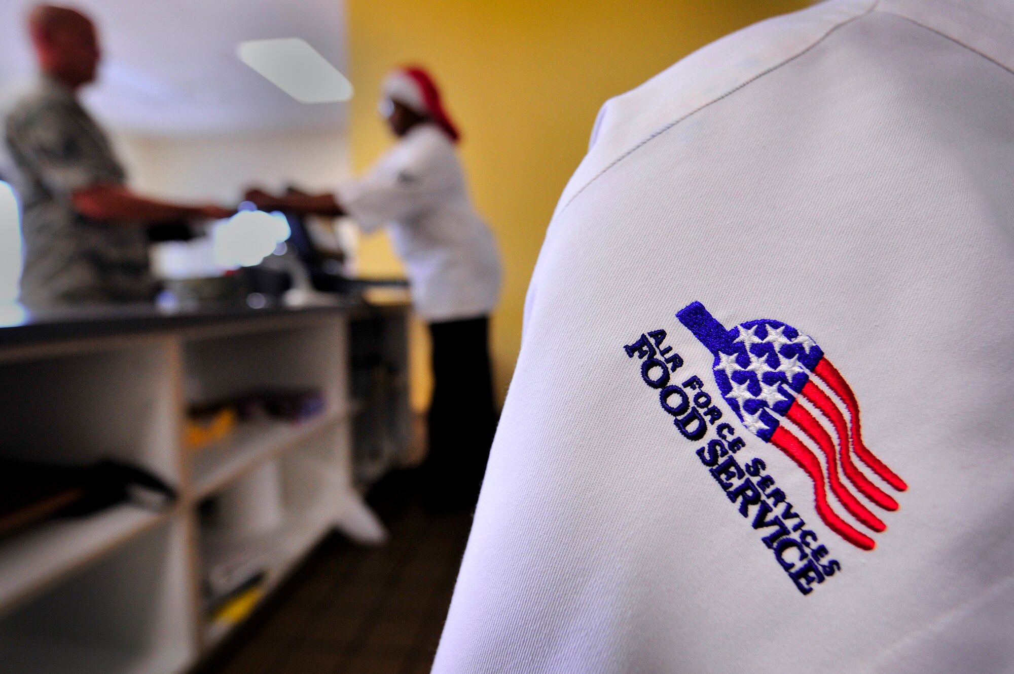 A U.S. Air Force Services food service coat hangs on a chair at the Afterburner Grill while Airmen assigned to the 20th Force Support Squadron help customers during a lunch hour rush, July 20, 2012, Shaw Air Force Base, S.C.  Air Force Food Service Specialists assigned to the 20th Force Support Squadron wear the food service symbol depicted in this photo while on duty. (U.S. Air Force photo by Senior Airman Kenny Holston/Released)