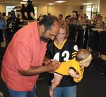 NFL Hall of Famer and four-time Super Bowl champion Franco Harris autographs the outfit of eight-month-old Matilyn Armenta as mother Savanna Armenta looks on July 12 at the Joint Base San Antonio-Lackland Military Clothing Store. (U.S. Air Force photo/Larry Kishur)