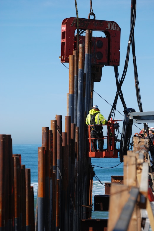 Working on a platform suspended above the Pacific Ocean, a worker for Granite Construction Company supervises as a crane nearby drives pilings during construction of Mexico-United States border fence.  