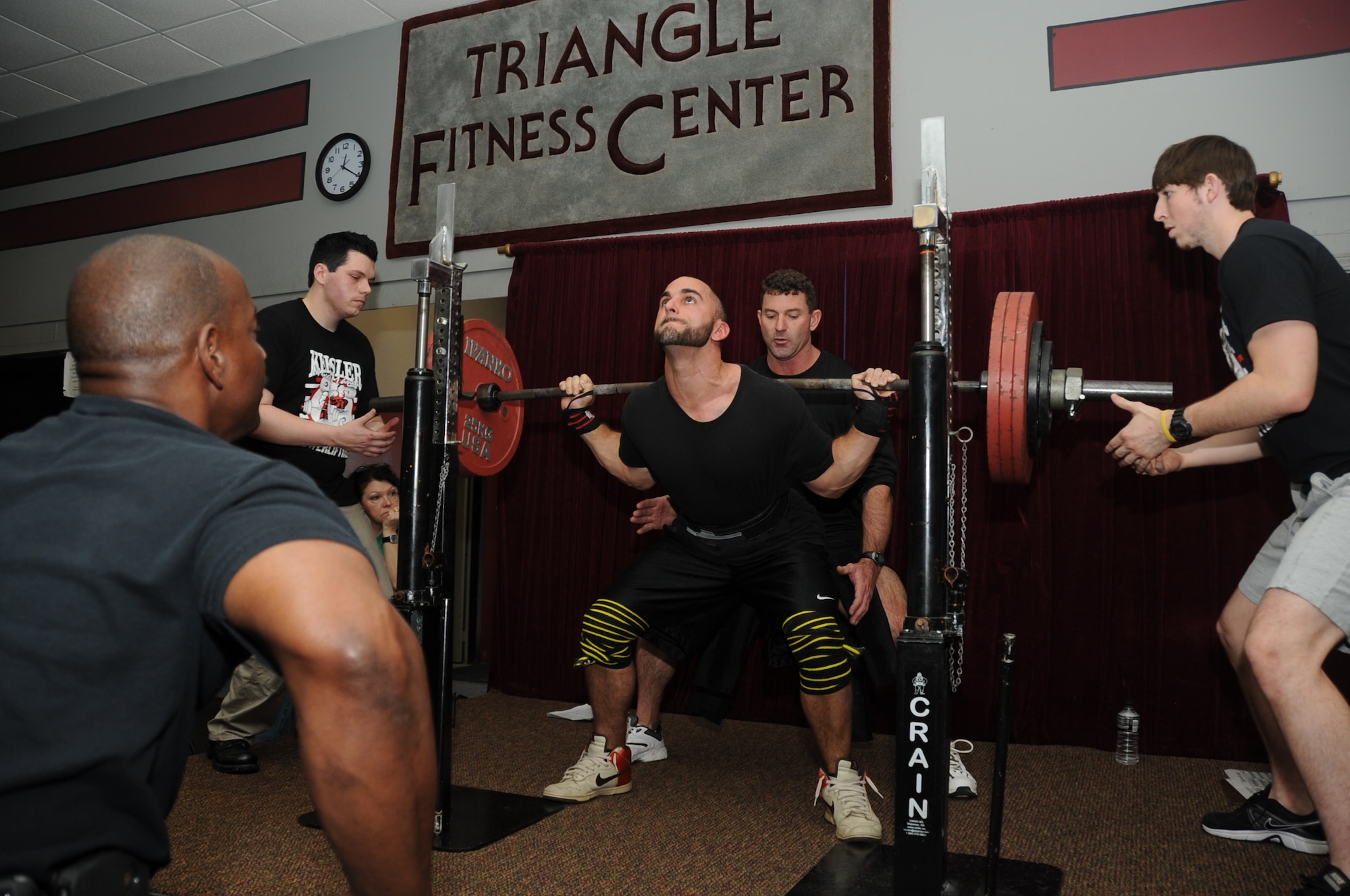 Al Watkins, 81st Infrastructure Division, judges the squatting competition as Justin Trott takes his turn squatting 425 pounds while triangle fitness staffers Lance Solari, Robert Reville and Kyle Kinsey spot him during the muscle mania competition March 24, 2012, Keesler Air Force Base, Miss.  (U.S. Air Force photo by Kemberly Groue)
