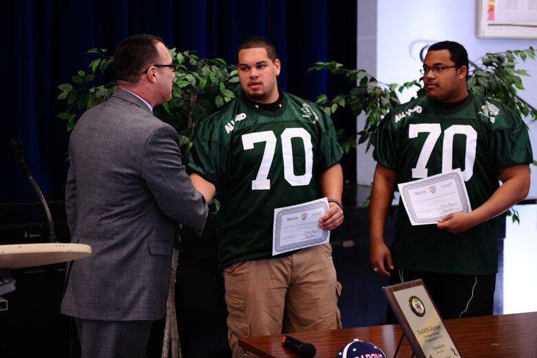 BITBURG ANNEX, Germany – Randy Plunkett, Military.com director of community and government outreach, shakes hands with Colton Engelmeier, congratulating him on his selection, as well as teammates Darian Billups and Matthew Flood, to the Military.com All-Department of Defense Team at Bitburg High School here March 27. The team is made up of 24 football players who are children of service members stationed throughout the world. Military.com is a military and veteran membership organization that connects service members, military families and veterans to all the benefits of service. The BHS football team members received a certificate and a team jersey, and will attend an upcoming invitation-only 2012 Football University Camp, taught by NFL coaches and players. The team members will find out at a later date which one of the 45 camps they will be invited to attend. (U.S. Air Force photo by Airman 1st Class Matthew B. Fredericks/Released)