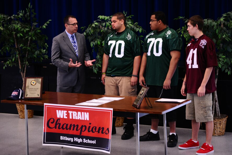 BITBURG ANNEX, Germany – Randy Plunkett, Military.com director of community and government outreach, congratulates, from left, Colton Engelmeier, Darian Billups and Matthew Flood on their selection to the Military.com All-Department of Defense Team at Bitburg High School here March 27. The team is made up of 24 football players who are children of service members stationed throughout the world. Military.com is a military and veteran membership organization that connects service members, military families and veterans to all the benefits of service. The BHS football team members received a certificate and a team jersey, and will attend an upcoming invitation-only 2012 Football University Camp, taught by NFL coaches and players. The team members will find out at a later date which one of the 45 camps they will be invited to attend. (U.S. Air Force photo by Airman 1st Class Matthew B. Fredericks/Released)
