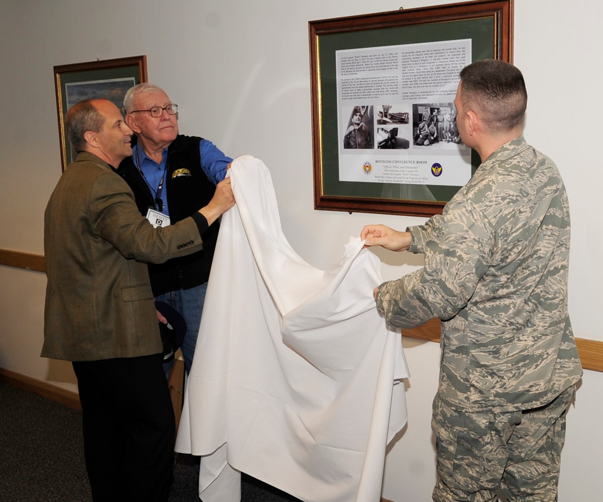 RAF MILDENHALL, England – Col. Joseph Rushlau, right, 100th Maintenance Group commander, unveils a framed poster of Maj. Eugene “Butch” Rovegno, during a dedication ceremony of the 100th MXG conference room, with the assistance of Richard Rovegno, left, and former master sergeant, crew chief and World War II veteran, Dewey Christopher May 8, 2012. Richard Rovegno is the grandson of the former World War II pilot and engineer who the 100th MXG conference room has been dedicated to. (U.S. Air Force photo/Senior Airman Ethan Morgan)