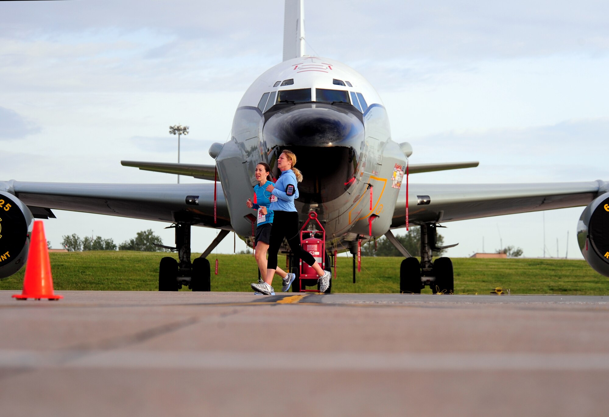The 55th Wing flagship RC-135V/W Rivet Joint sits behind the Offutt Field House as runners pass by during the Bellevue-Offutt Runway Run held at Offutt Air Force Base, Neb., May 13. Nearly 200 runners are guided down the runway every Mother's Day while they take part in a unique, seven mile run that consists of running down the Offutt runway and around various landmarks located at the base.  (U.S. Air Force Photo by Josh Plueger)