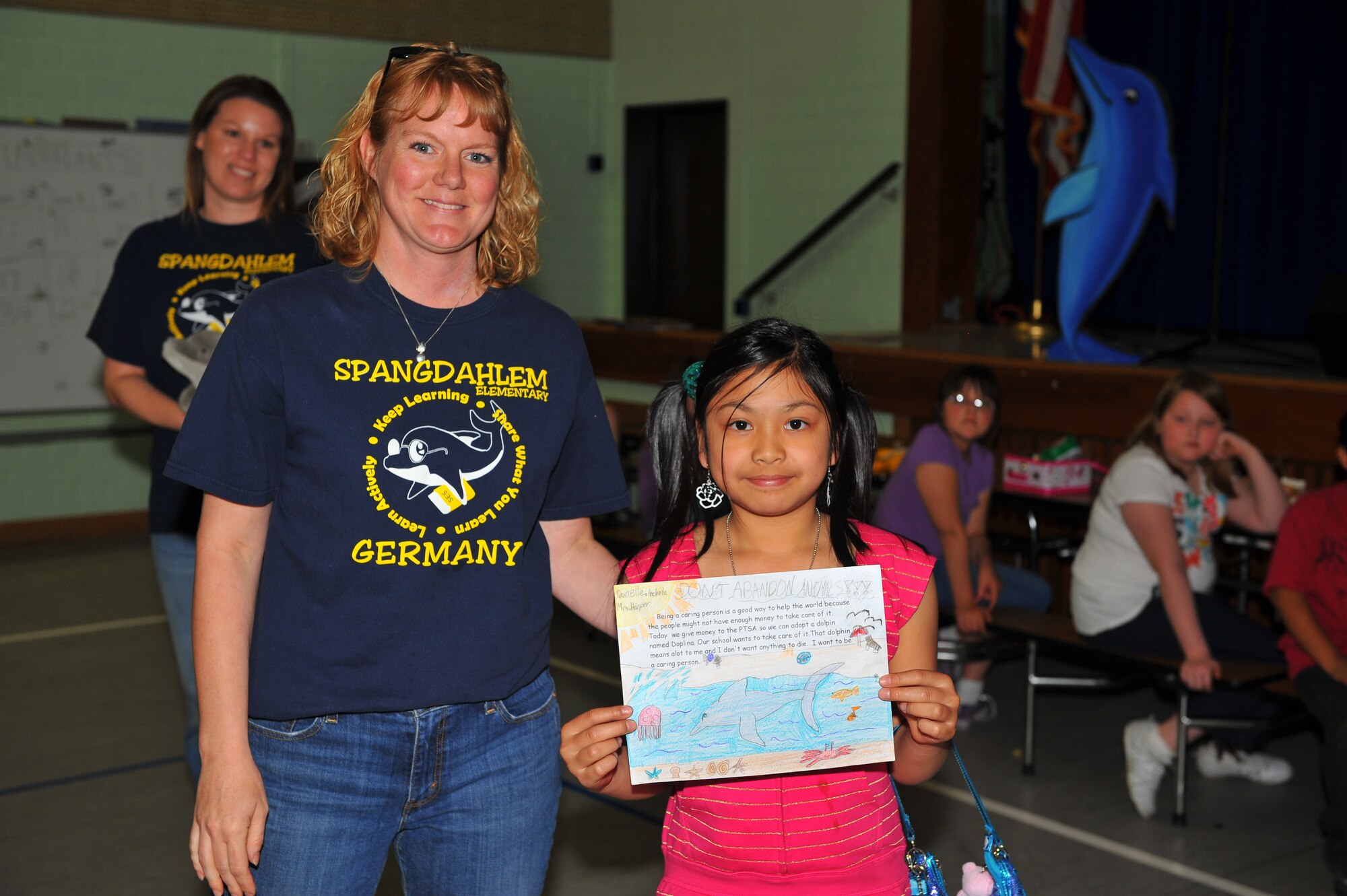 SPANGDAHLEM AIR BASE, Germany – Amy Finley, left, Spangdahlem Elementary School Parent Teacher Student Association president, and Danielle Ancheta, third grade student and daughter of Tech. Sgt. Ryan Ancheta, pose for a photograph with Ancheta’s first-place dolphin article during the initial presentation of Delfina’s adoption certificate inside the SES cafeteria here May 25. Delfina is a wild bottlenose dolphin the school adopted for one year through the World Wildlife Fund with help from the SES PTSA. The WWF uses the donations to help protect and improve living conditions for wildlife species around the globe. (U.S. Air Force photo by Airman 1st Class Dillon Davis/Released)