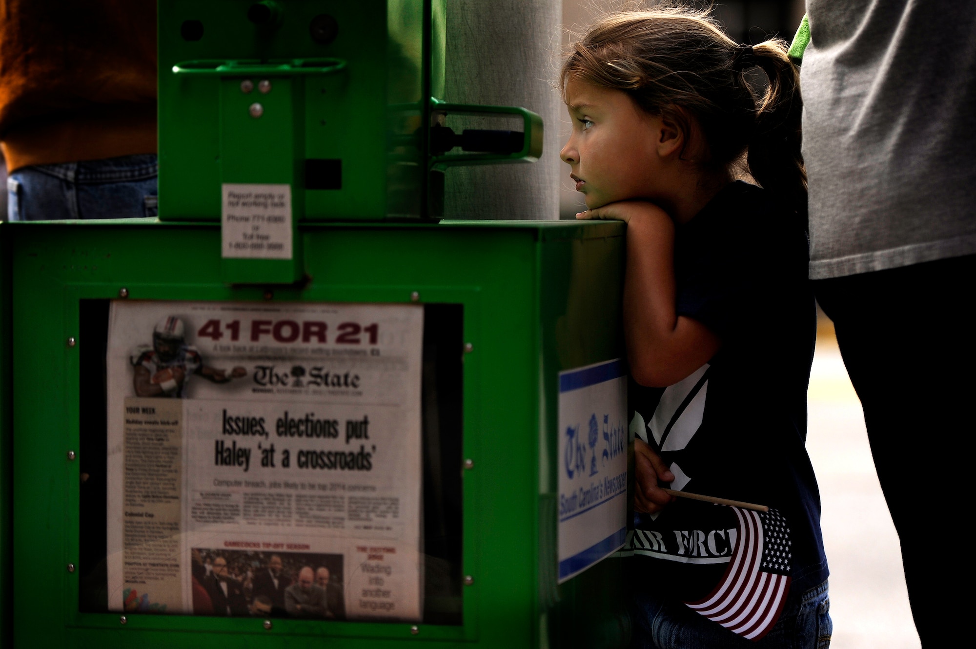 The daughter of a U.S. Air Force member holds an American flag as she leans against a newspaper stand while attending a Veterans Day Parade in Columbia, S.C., Nov. 12, 2012, just days after the United States presidential election. The newspaper stand contains The State’s most recent publication headlining South Carolina Governor Nikki Haley’s having reached a crossroads’ as a result of the Obama reelection. According to examiner.com Haley stated, “I will work together with President Obama wherever I can for the betterment of our state and country.” During Veterans Day Haley released a statement saying “May god continue to bless our veterans and their families and keep them strong and may he continue to bless the United States of America.” (U.S. Air Force photo by Staff Sgt. Kenny Holston/Released)  