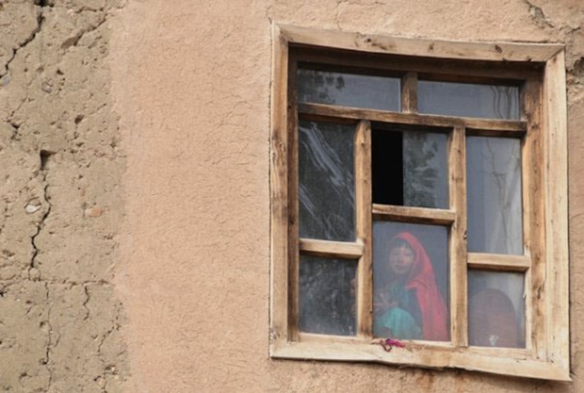An Afghan girl smiles behind a window in Ghazni province, Afghanistan in 20009. Then-Tech. Sgt. Rebecca Corey was documenting the work of the provincial reconstruction team in the region, when she photographed the child standing in the window observing her team. (U.S. Air Force photo/Master Sgt. Rebecca Corey)