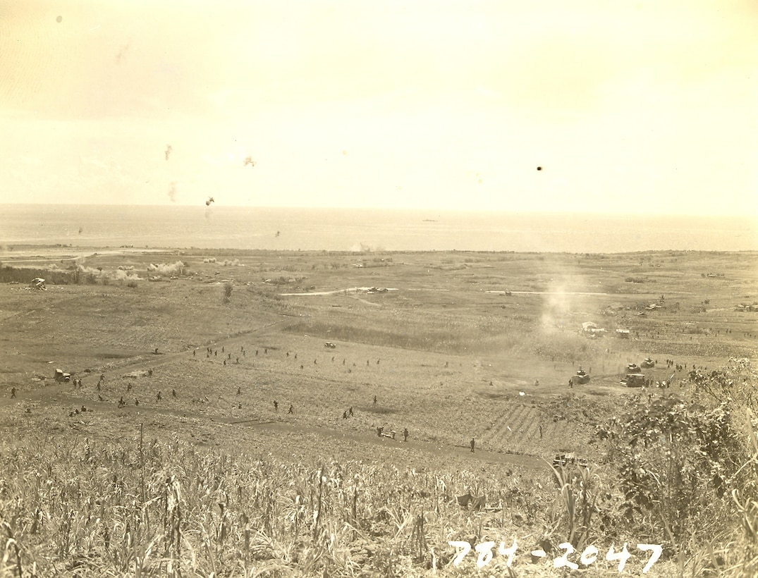 "Attack moves to the northern end of Saipan. The island was secured the same day. An airfield is on upper left & just to the right of it are puffs of smoke from artillery hits. I saw this also from this height - it was quite a scene."