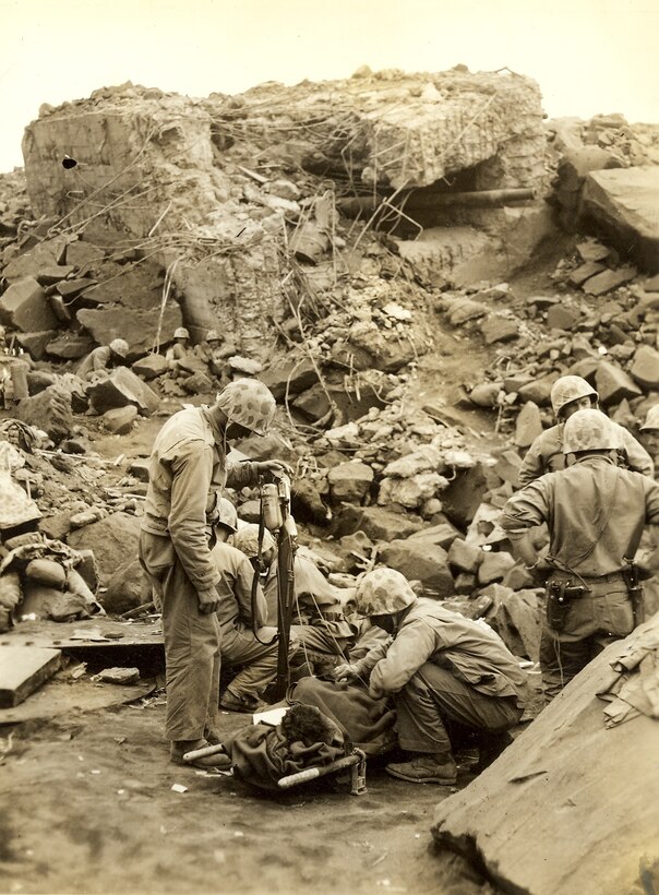 Corpsmen are providing medical treatment to Marines in foreground, Marines are dug in behind a demolished artillery bunker.