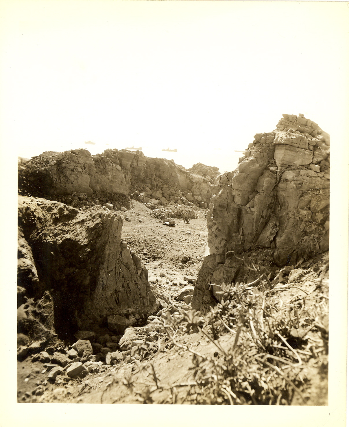 Two men walk through rock quarry, U.S. Naval Forces are stationed off shore in the background.