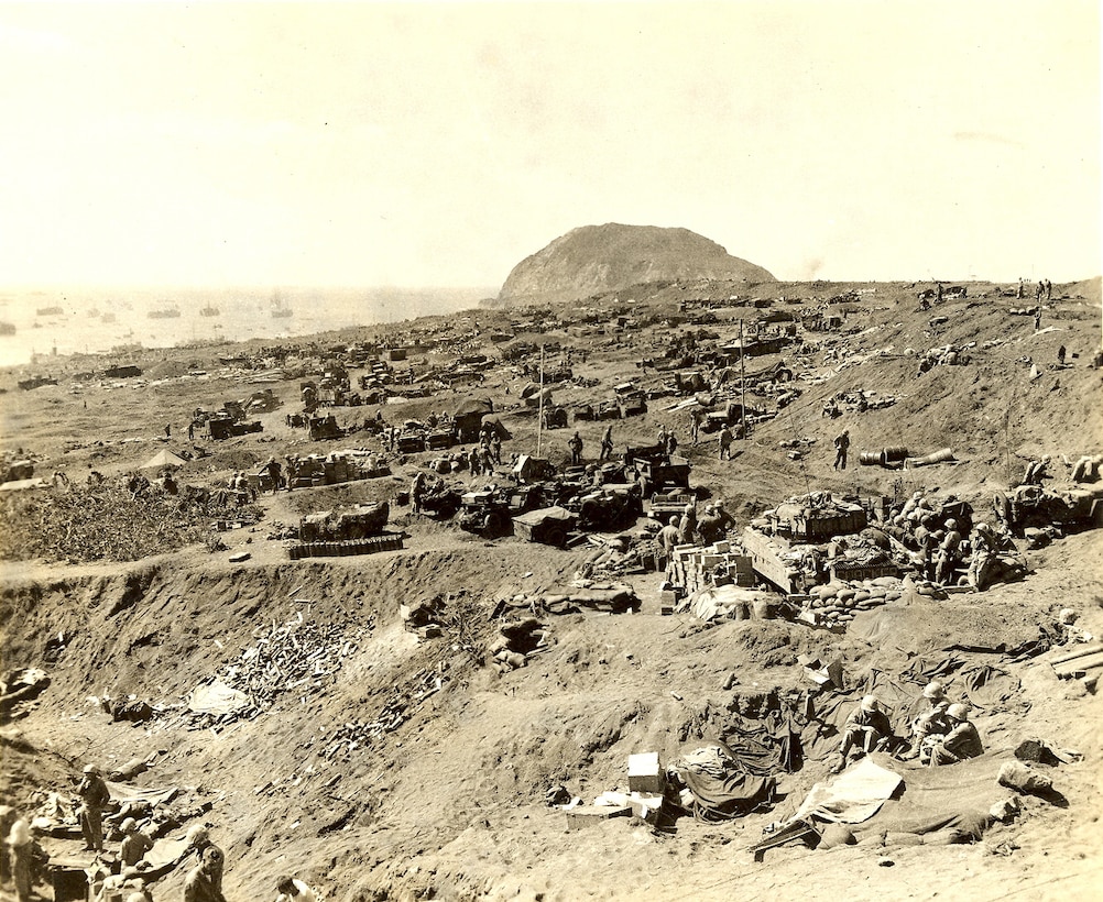 Two men walk through rock quarry, U.S. Naval Forces are stationed off shore in the background.